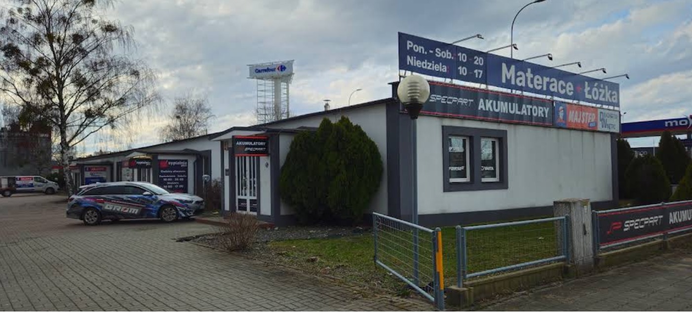 Budynek warsztatu samochodowego Specpart z reklamą akumulatorów i materacy, widoczny samochód z logo firmy i baner reklamowy, w tle maszt z logo Carrefour.