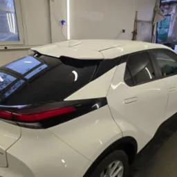 Biały samochód Toyota C-HR Hybrid po umyciu, widoczne krople wody na szybie, zaparkowany w hali z oświetleniem warsztatowym.