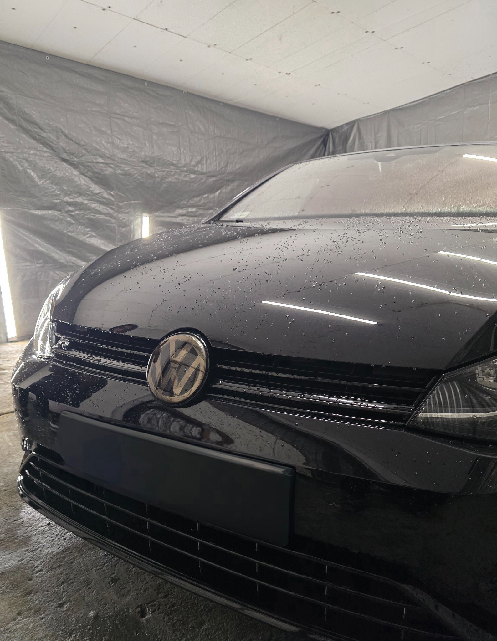 Czarny samochód marki Volkswagen, pokryty kroplami wody, po umyciu w myjni, widok z przodu na logo i atrapę chłodnicy, w tle szara plandeka i oświetlenie warsztatowe.