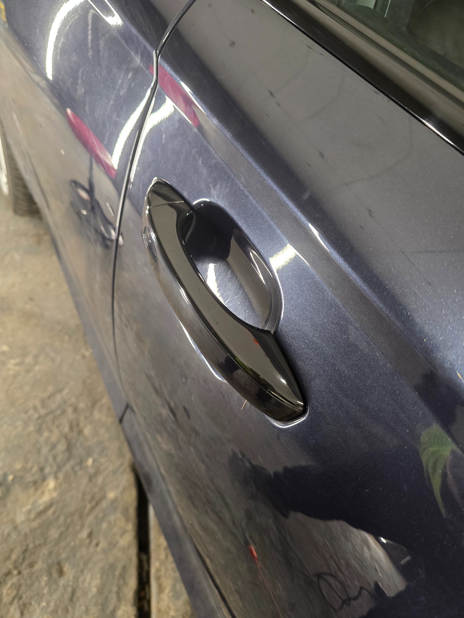 Błyszcząca, czarna klamka na ciemnoszarym samochodzie z widocznymi smugami i odbiciami na lakierze.