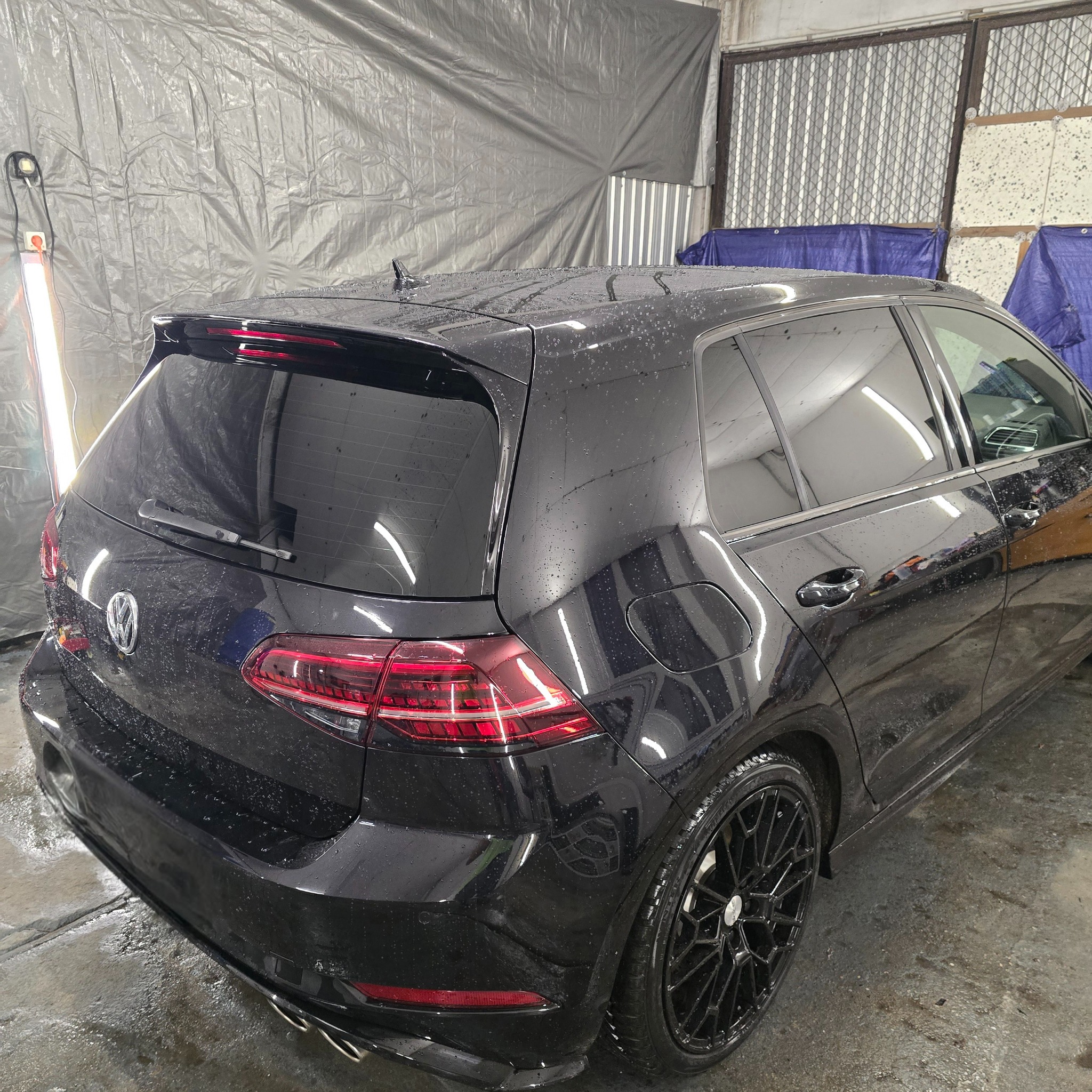 Czarny Volkswagen Golf po myciu, z kroplami wody na lakierze, widoczne tylne światła i felgi z czarnymi obręczami, w tle szara plandeka i oświetlenie warsztatowe.
