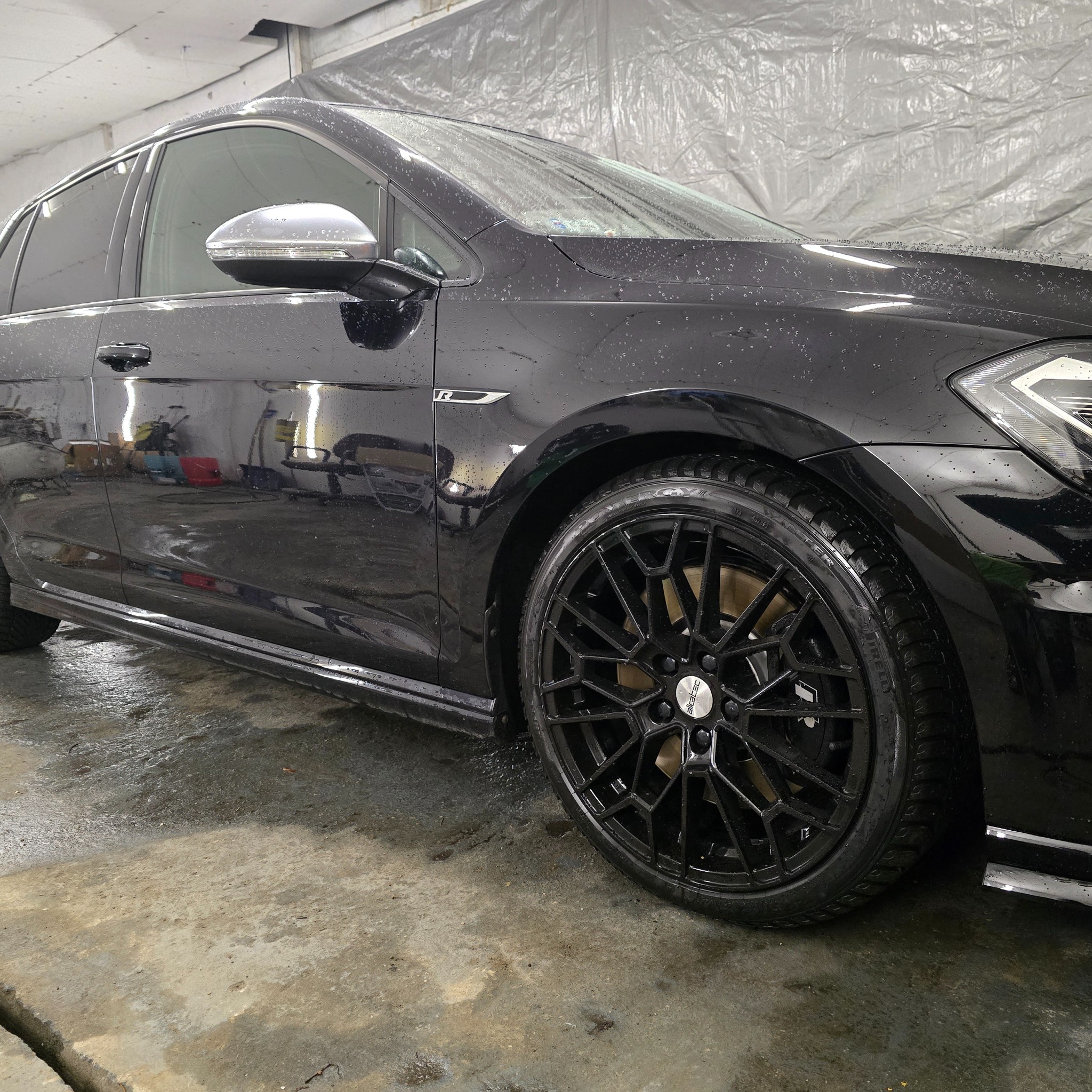 Czarny samochód typu kombi, mokry po umyciu, z widocznymi kroplami wody na karoserii i felgach z wzorem w kształcie gwiazdy, stojący w myjni.