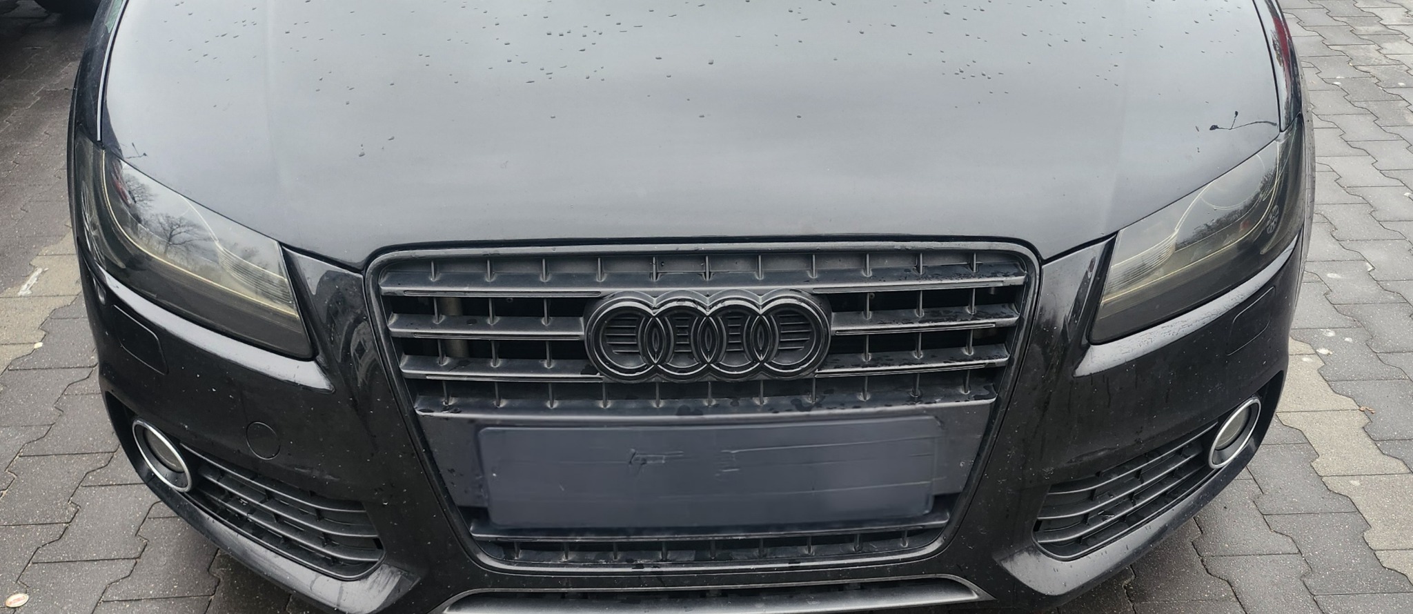 Przód czarnego samochodu marki Audi z widocznymi śladami deszczu na masce i reflektorach.