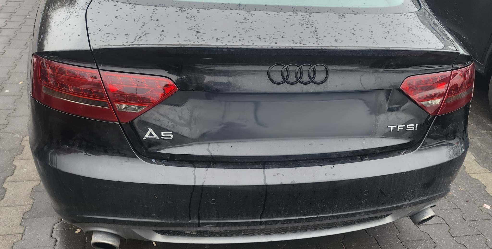Tył czarnego samochodu Audi A5 TFSI z widocznymi kroplami wody na karoserii, zaparkowanego na chodniku z kostki brukowej.