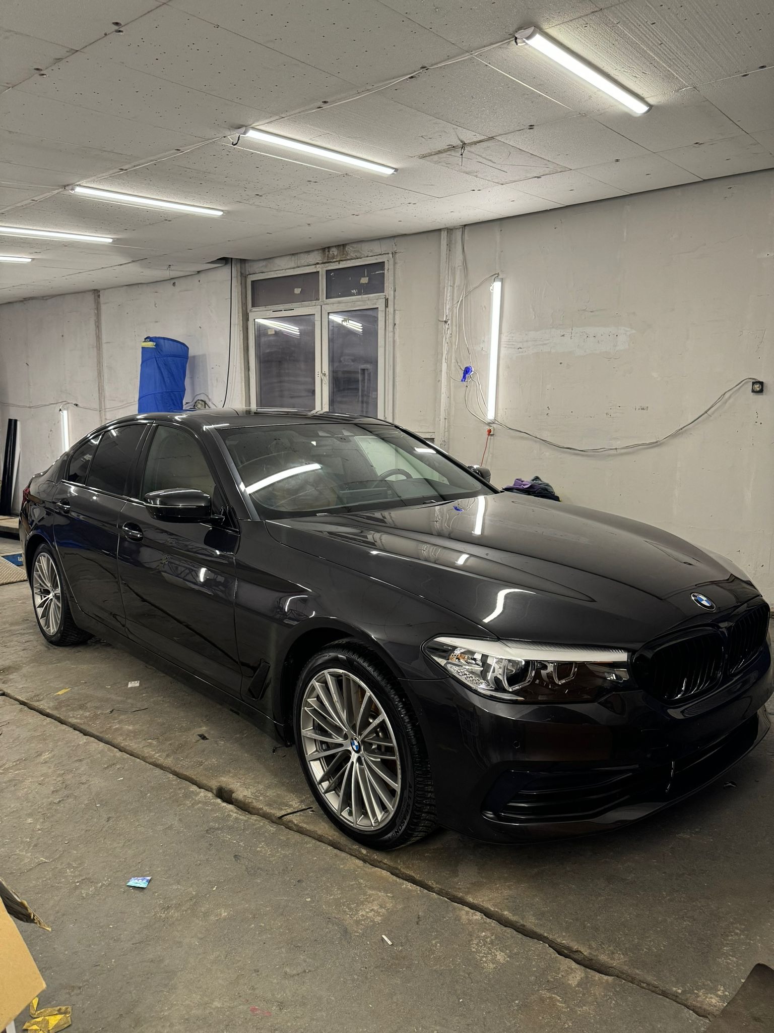 Ciemny sedan BMW po przyciemnianiu szyb, widoczne ciemne szyby boczne i tylna, zaparkowany w warsztacie z betonową podłogą i oświetleniem jarzeniowym.