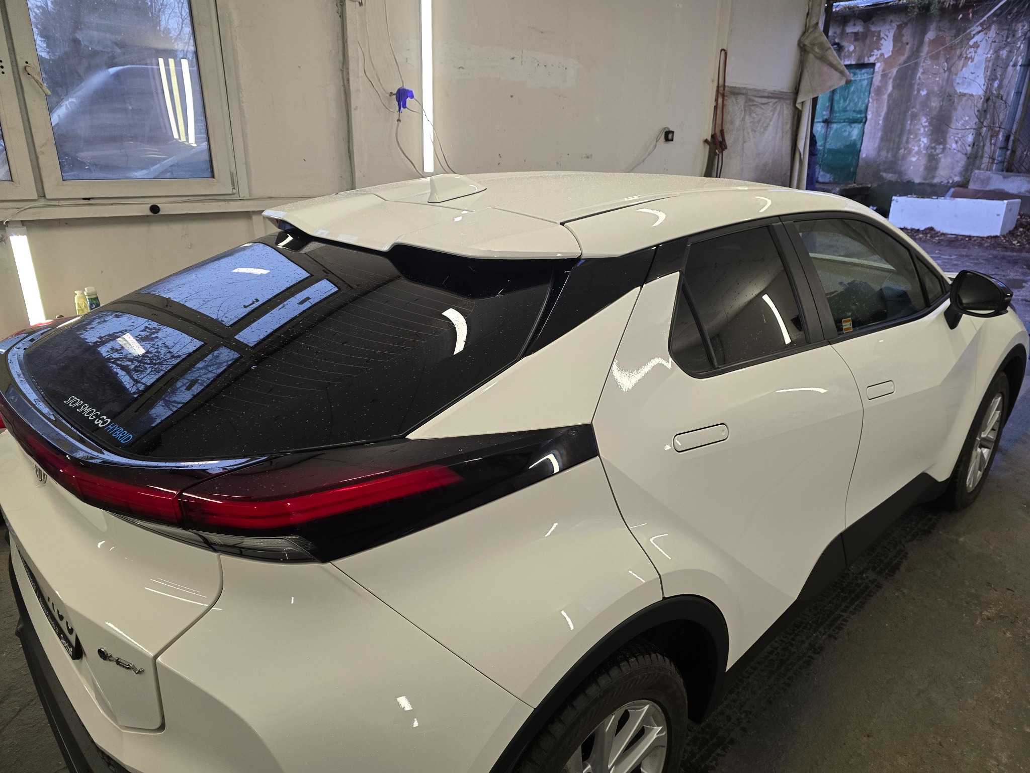 Biały samochód Toyota C-HR Hybrid po umyciu, widoczne krople wody na szybie, zaparkowany w hali z oświetleniem warsztatowym.