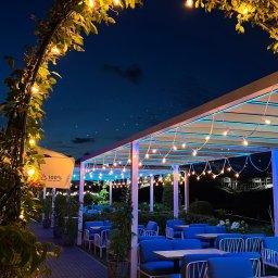 SMART EV - Oświetlony ogródek restauracyjny nocą, z roślinnym łukiem i girlandami żarówek, niebieskie podświetlenie pod zadaszeniem, nowoczesne meble ogrodowe w odcieniach błękitu.