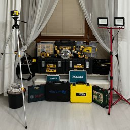 SMART EV - Zestaw narzędzi elektrycznych różnych marek (DeWalt, Makita, Bosch, Högert, Nivel System, Stanley) ułożonych przed oknem z zasłonami i oświetlaczami LED na statywach, obejmujący m.in. wiertarkę...