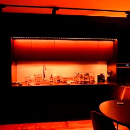 SMART EV - Nowoczesna kuchnia w Warszawie z podświetleniem LED w kolorze pomarańczowym, oświetlającym blat i szafki wiszące. Na blacie widoczne akcesoria kuchenne i dekoracje. Na podłodze leżą narzędzia...