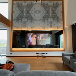 SMART EV - Salon z dużą marmurową ścianą za telewizorem, podświetlonymi listwami LED i nowoczesną lampą wiszącą, widok zza szarej sofy.