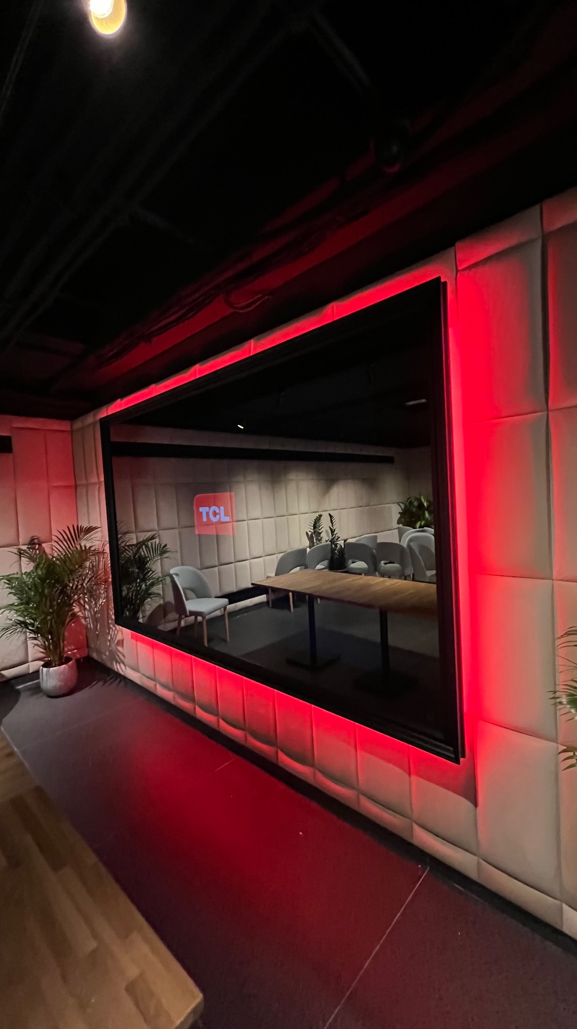 Nowoczesna sala konferencyjna z podświetleniem LED na obrzeżach, widoczna przez duże okno. Wnętrze wyłożone panelami ściennymi, wyposażone w stół konferencyjny i krzesła. Widoczny logotyp TCL...