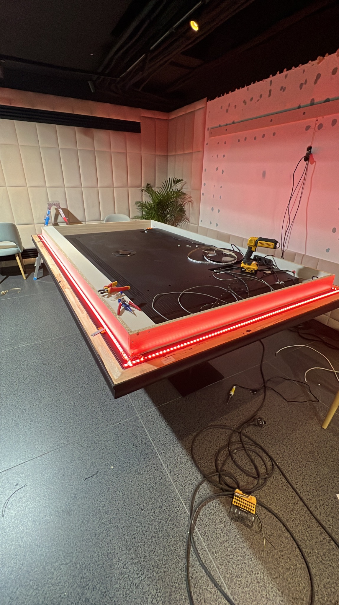 Montaż oświetlenia LED w ramie stołu, widoczne narzędzia, przewody, wiertarka akumulatorowa DeWalt, szczypce, czerwone światło odbija się od ściany, w tle tapicerowana ściana.