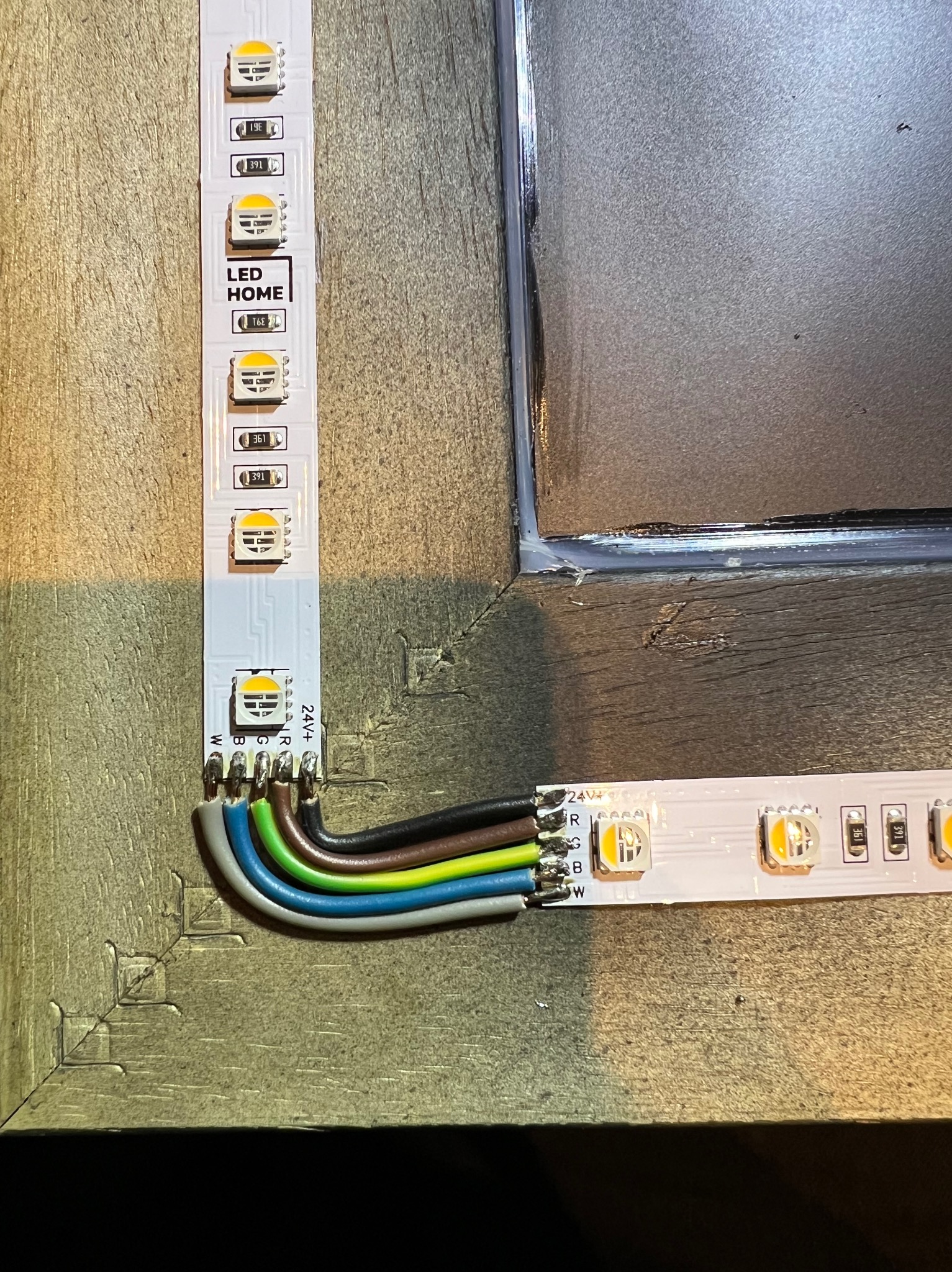 Zbliżenie na podłączoną taśmę LED RGB z widocznymi przewodami (szary, niebieski, zielony, brązowy i czarny) oraz fragmentem ramy lustra z silikonem, na tle drewnopodobnej powierzchni.