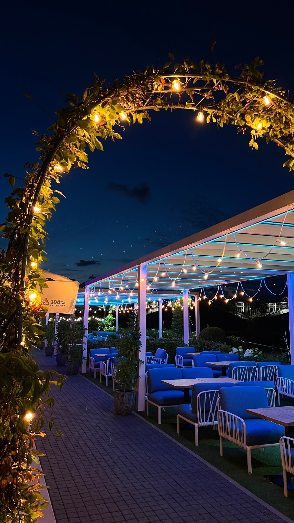 Oświetlony ogródek restauracyjny nocą, z roślinnym łukiem i girlandami żarówek, niebieskie podświetlenie pod zadaszeniem, nowoczesne meble ogrodowe w odcieniach błękitu.