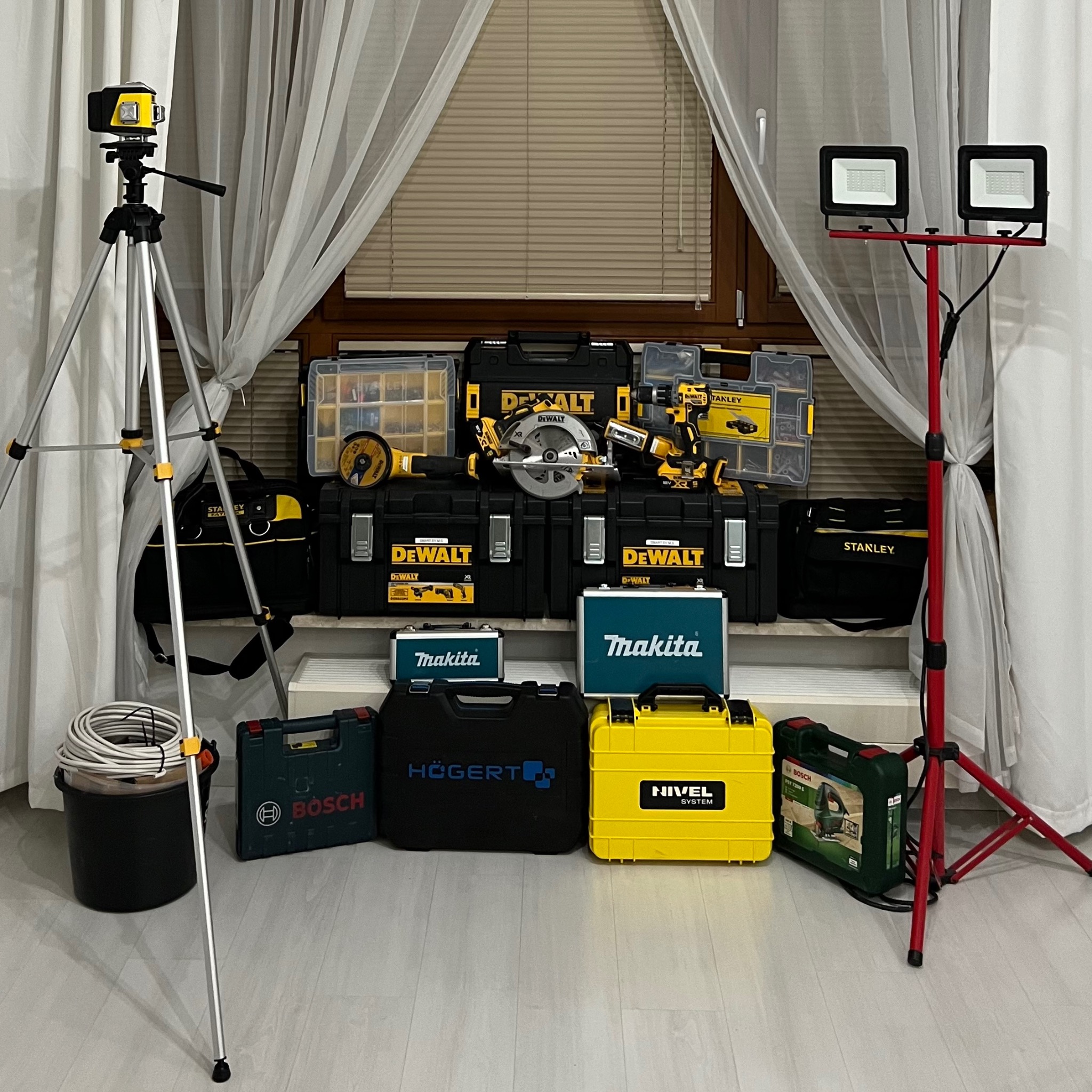 Zestaw narzędzi elektrycznych różnych marek (DeWalt, Makita, Bosch, Högert, Nivel System, Stanley) ułożonych przed oknem z zasłonami i oświetlaczami LED na statywach, obejmujący m.in. wiertarkę...