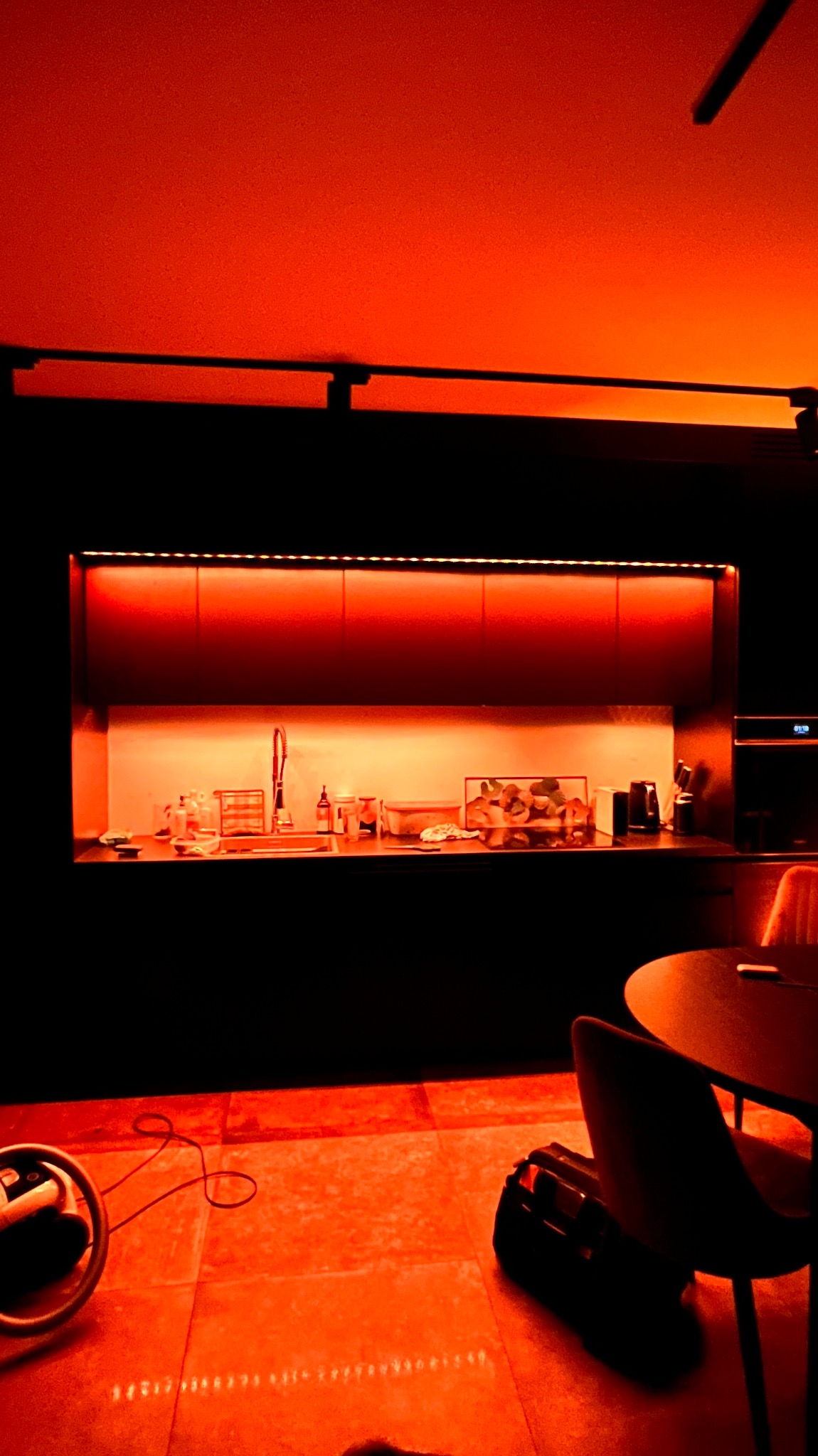 Nowoczesna kuchnia w Warszawie z podświetleniem LED w kolorze pomarańczowym, oświetlającym blat i szafki wiszące. Na blacie widoczne akcesoria kuchenne i dekoracje. Na podłodze leżą narzędzia...