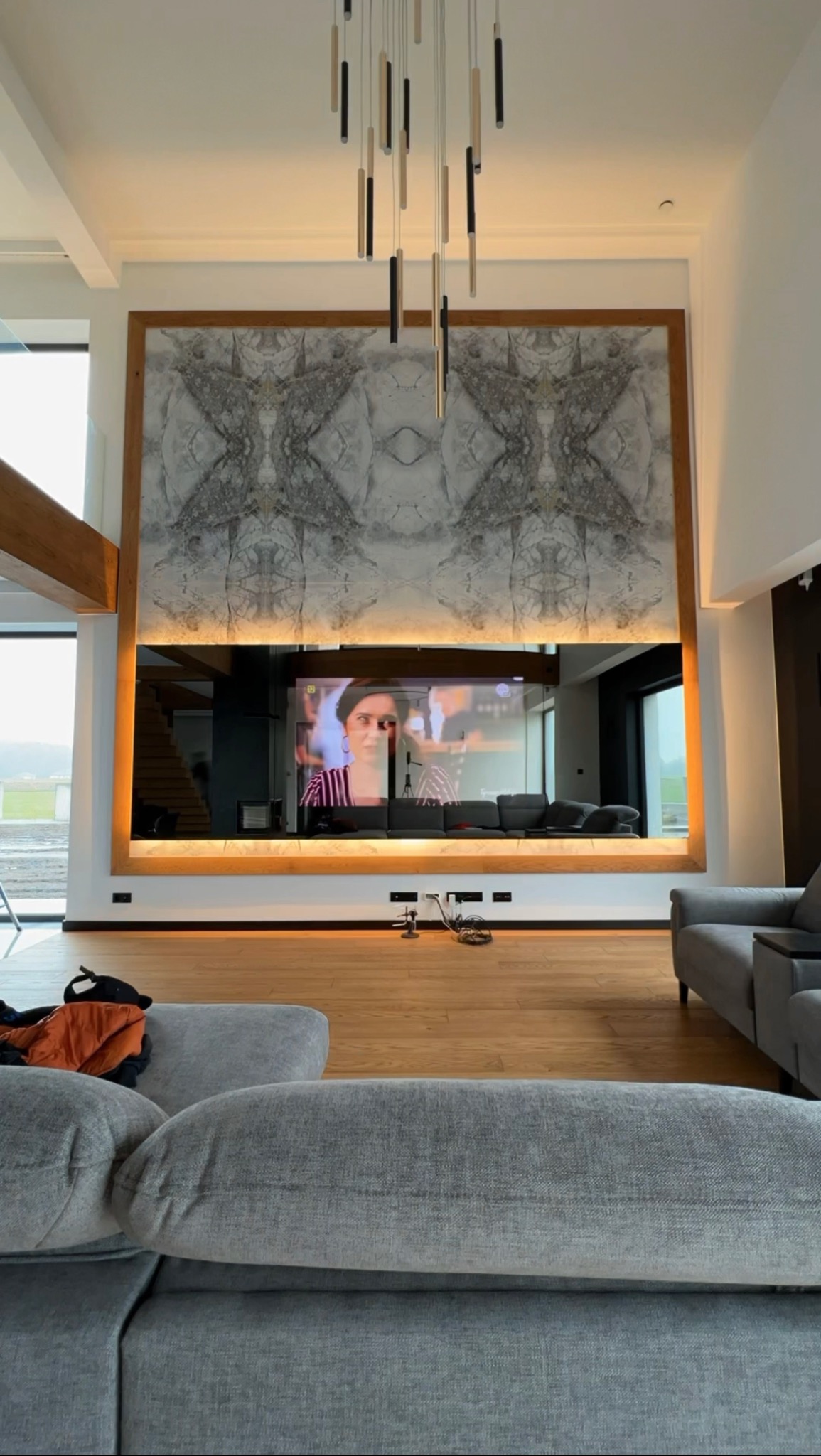Salon z dużą marmurową ścianą za telewizorem, podświetlonymi listwami LED i nowoczesną lampą wiszącą, widok zza szarej sofy.