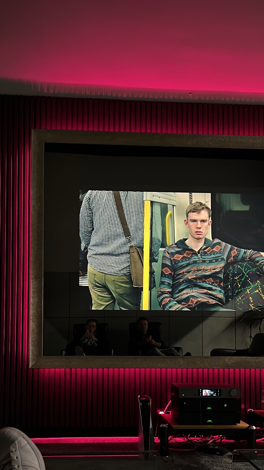 Kino domowe z dużym ekranem wyświetlającym film, podświetlenie LED w kolorze fuksji oświetla ścianę z pionowymi panelami, widoczny sprzęt audio i konsola Playstation 5.