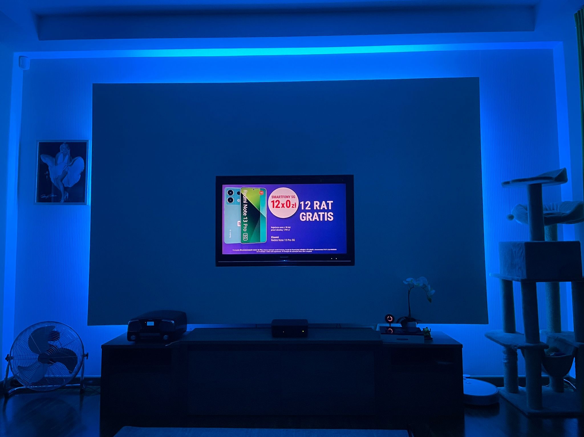 Salon z telewizorem wyświetlającym reklamę smartfona Xiaomi Redmi Note 13 Pro 5G, oświetlony niebieskim światłem LED, z widocznym wentylatorem i drapakiem dla kota.