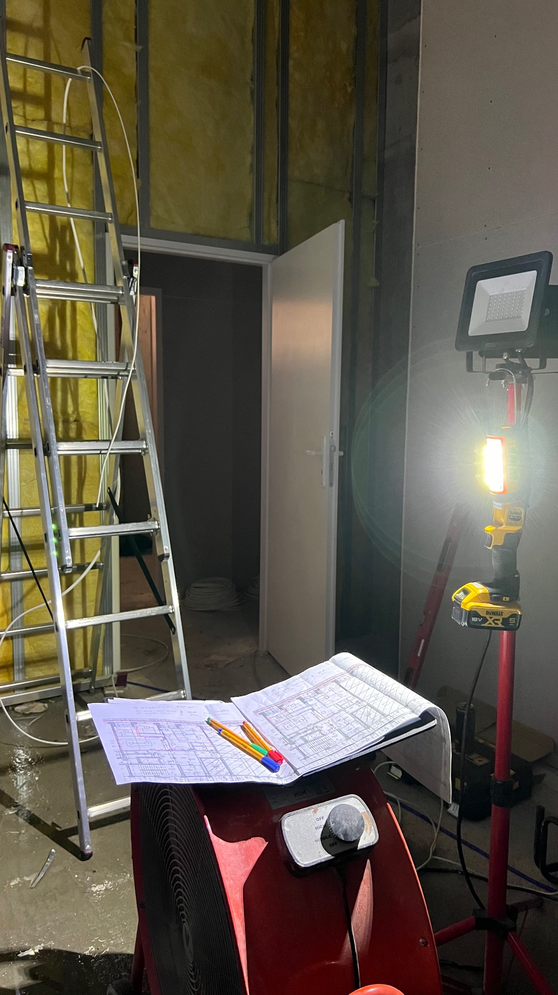 Wnętrze w trakcie prac wykończeniowych: drabina oparta o ścianę z izolacją, rozłożone plany na nagrzewnicy, oświetlenie robocze.