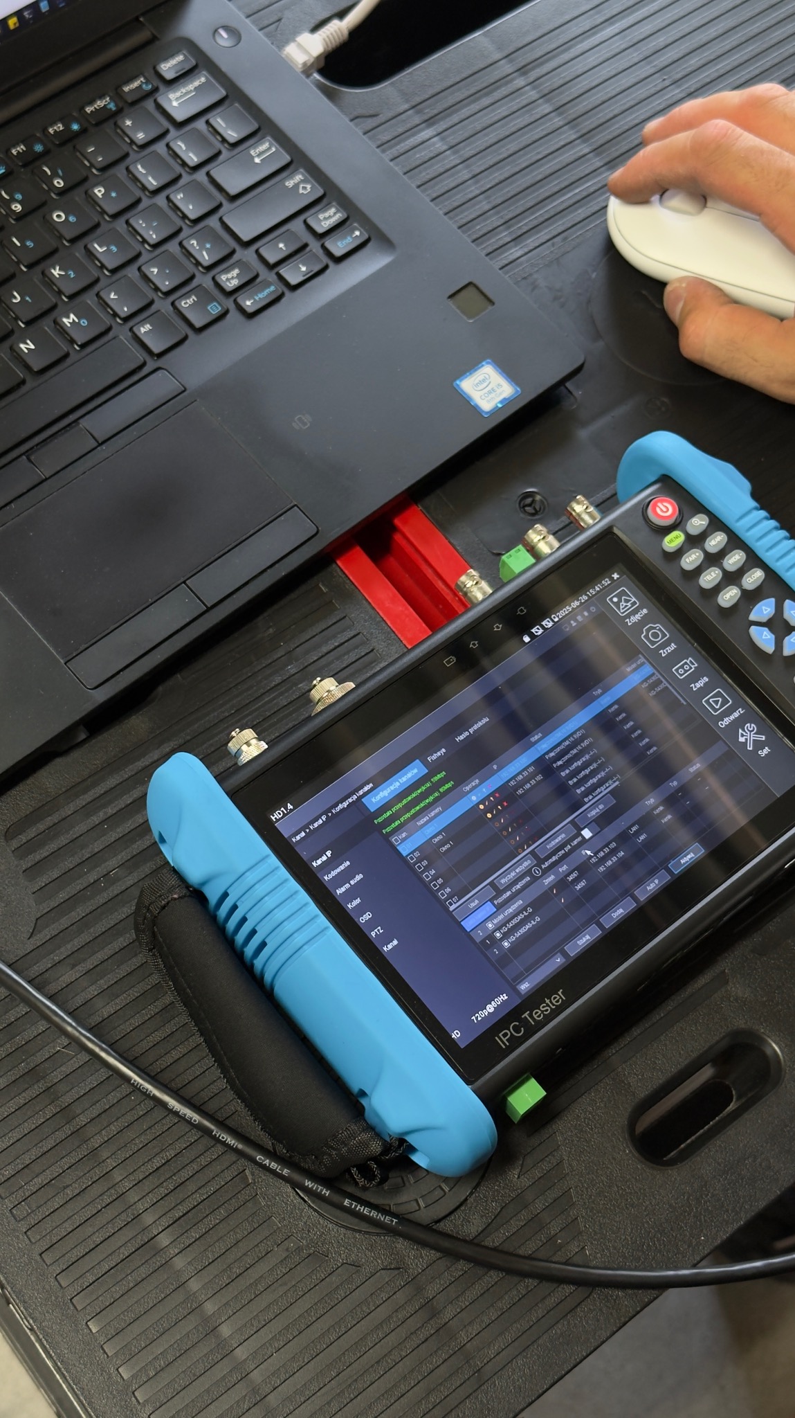 Tester IPC podłączony do laptopa, wyświetla konfigurację kanałów i ustawienia kamer. Kabel Ethernet i mysz komputerowa widoczne w kadrze.