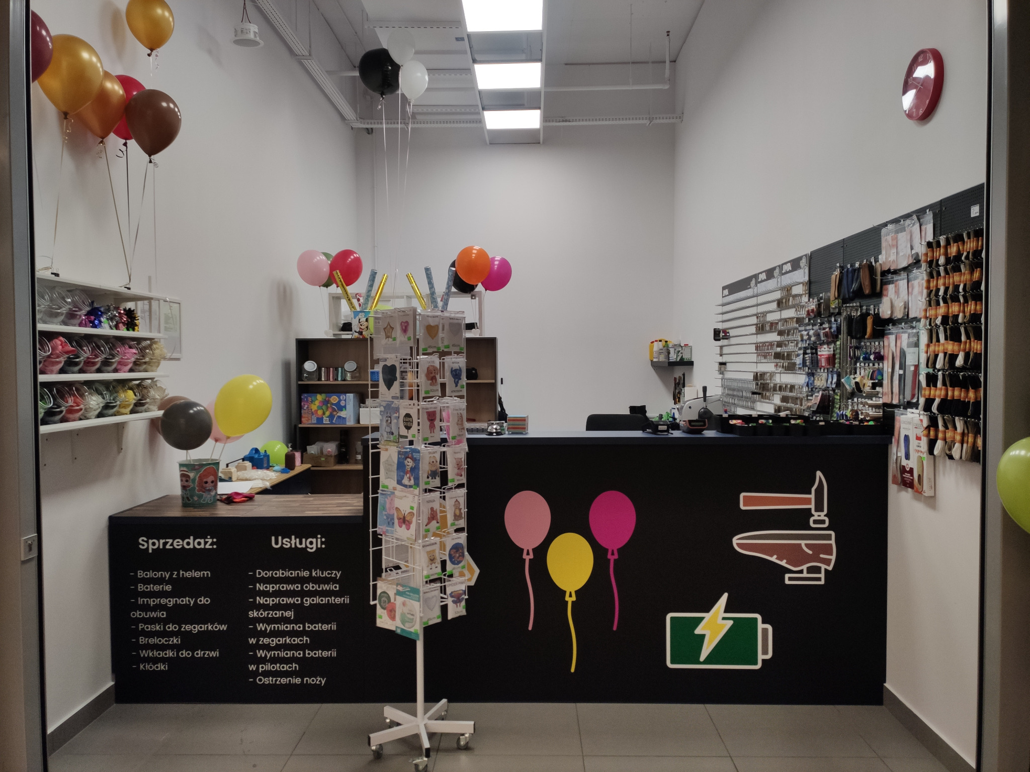 Wnętrze punktu usługowego z ladą, na której widoczne są informacje o sprzedaży i usługach, takie jak dorabianie kluczy, naprawa obuwia i wymiana baterii, dekoracje z balonów.