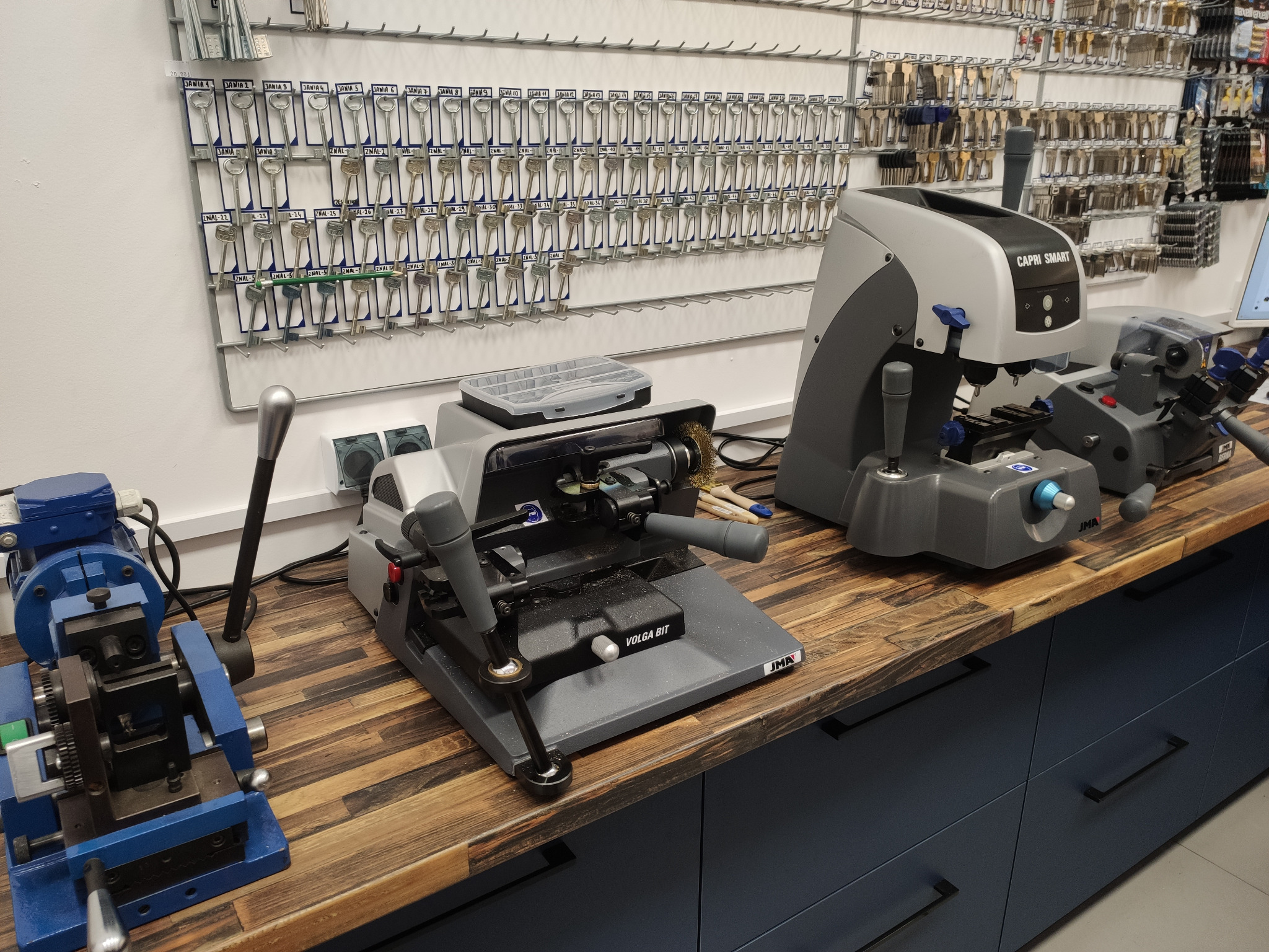 Warsztat z maszynami do kopiowania kluczy Capri Smart i Volga Bit, widok na ścianę z zawieszkami na klucze z opisami.