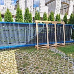 Gold Fence - Szare panele ogrodzeniowe z niebieską ramą, zabezpieczone folią, oparte o drewnianą paletę na trawniku z ażurowych płyt, w tle rząd wysokich tuj i budynek w stanie surowym.