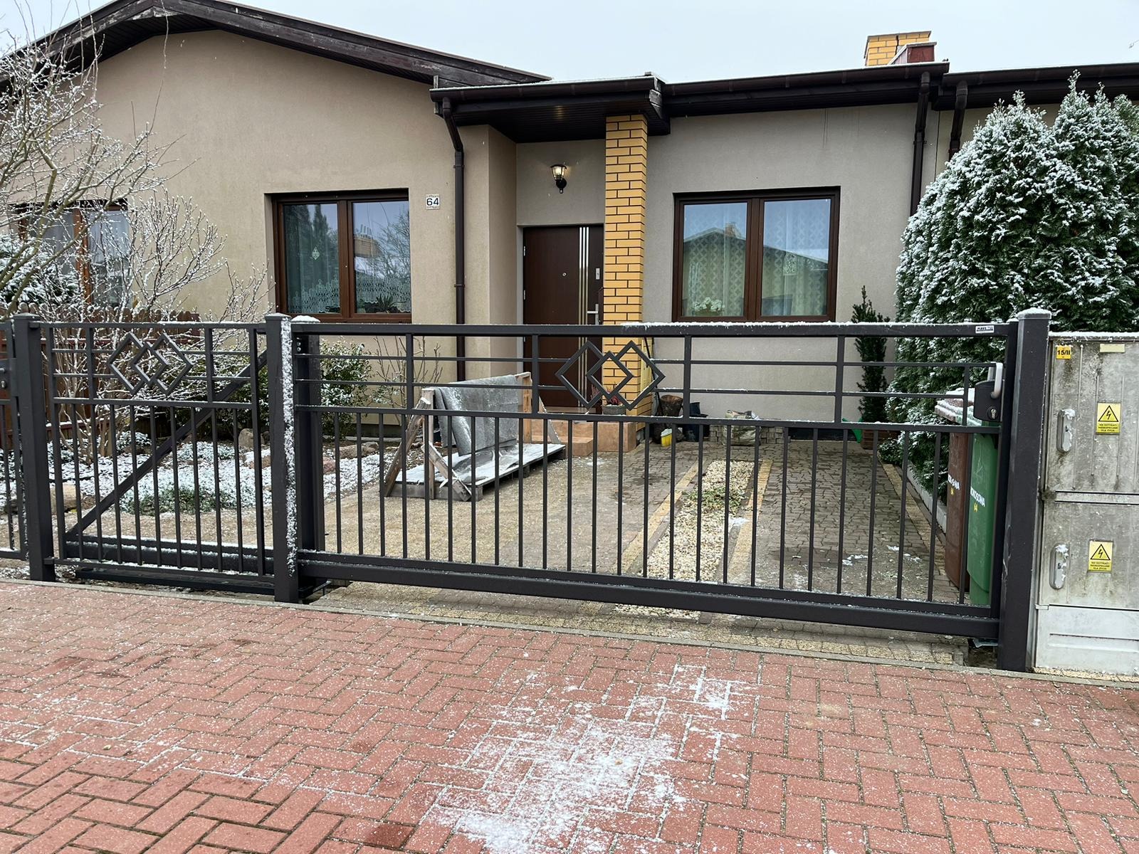 Przesuwna brama z metalowych prętów na posesji z kostki, z elementami dekoracyjnymi w górnej części, widoczny fragment domu z numerem 64, trawnik z krzewami pokryty śniegiem.
