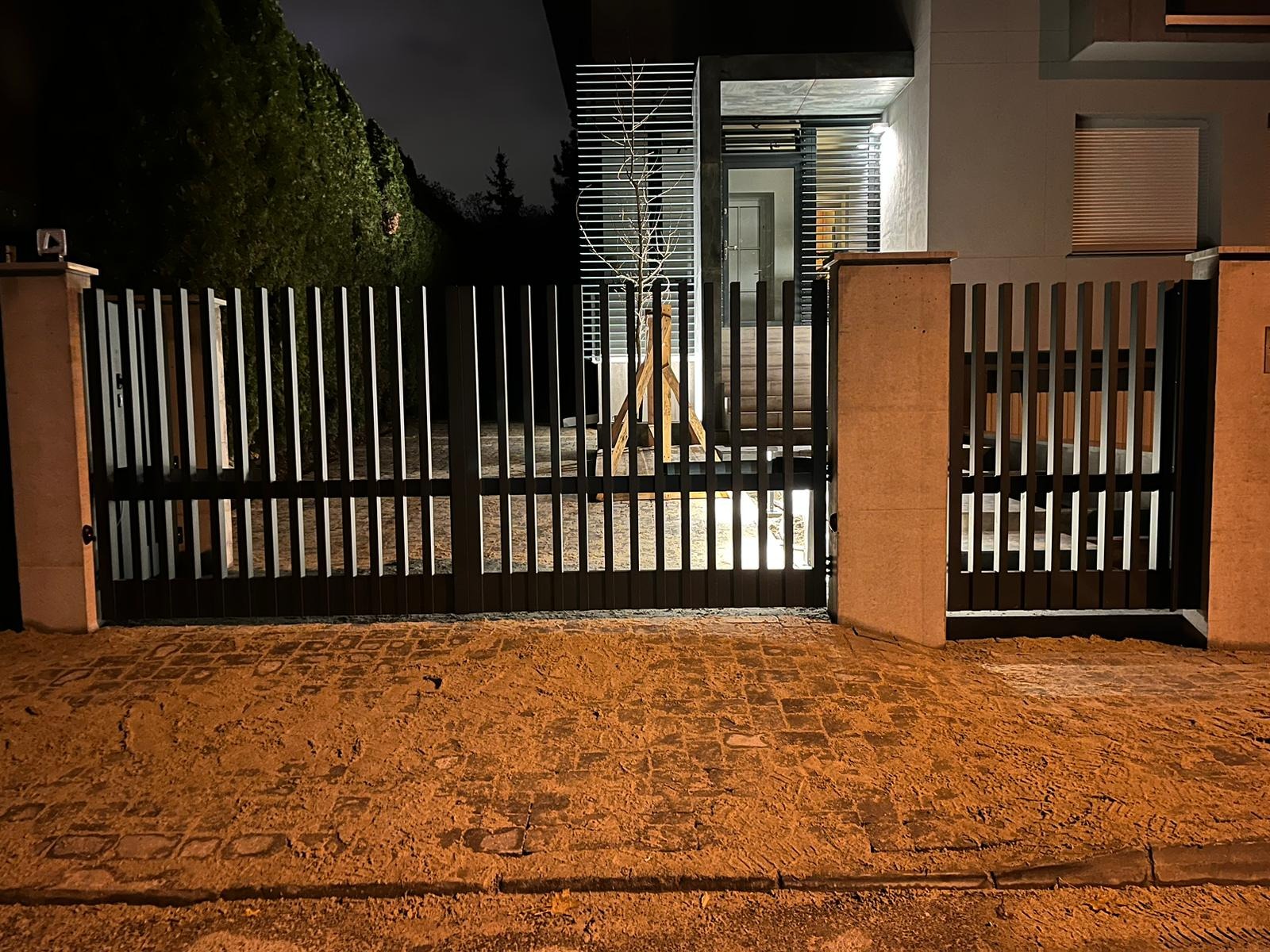 Nowoczesna, czarna brama wjazdowa z pionowych profili, oświetlona od dołu, z widocznym młodym drzewkiem wewnątrz posesji, sfotografowana nocą.