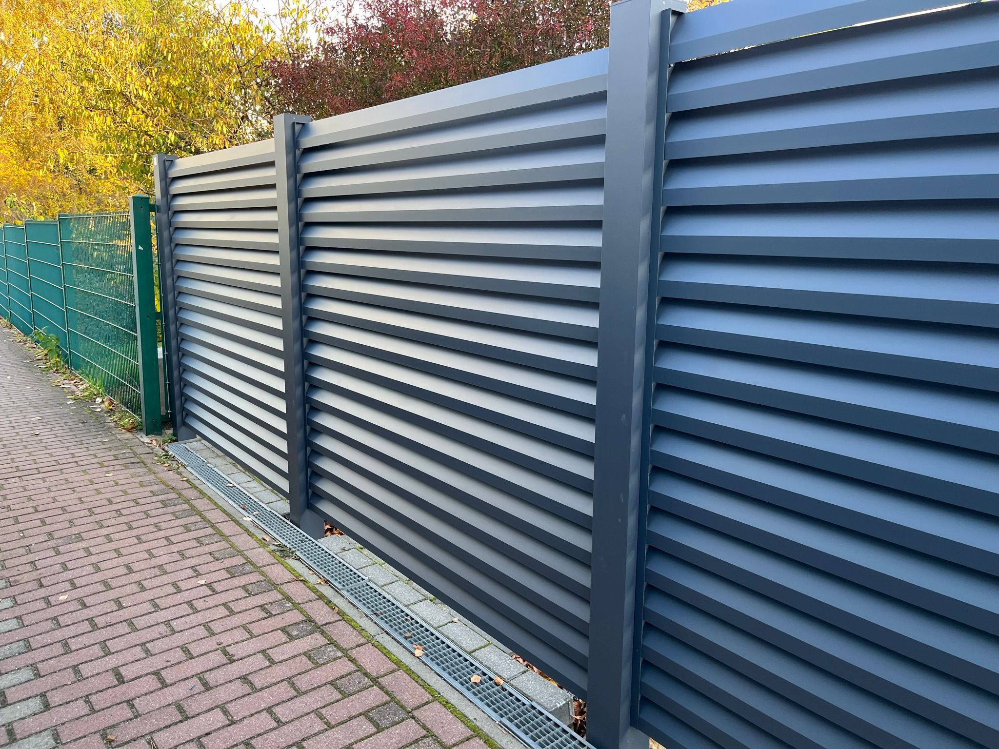 Nowoczesne, szare ogrodzenie lamelowe z metalu, widok z boku na tle zielonej siatki i jesiennych drzew, z odpływem liniowym przy kostce brukowej.