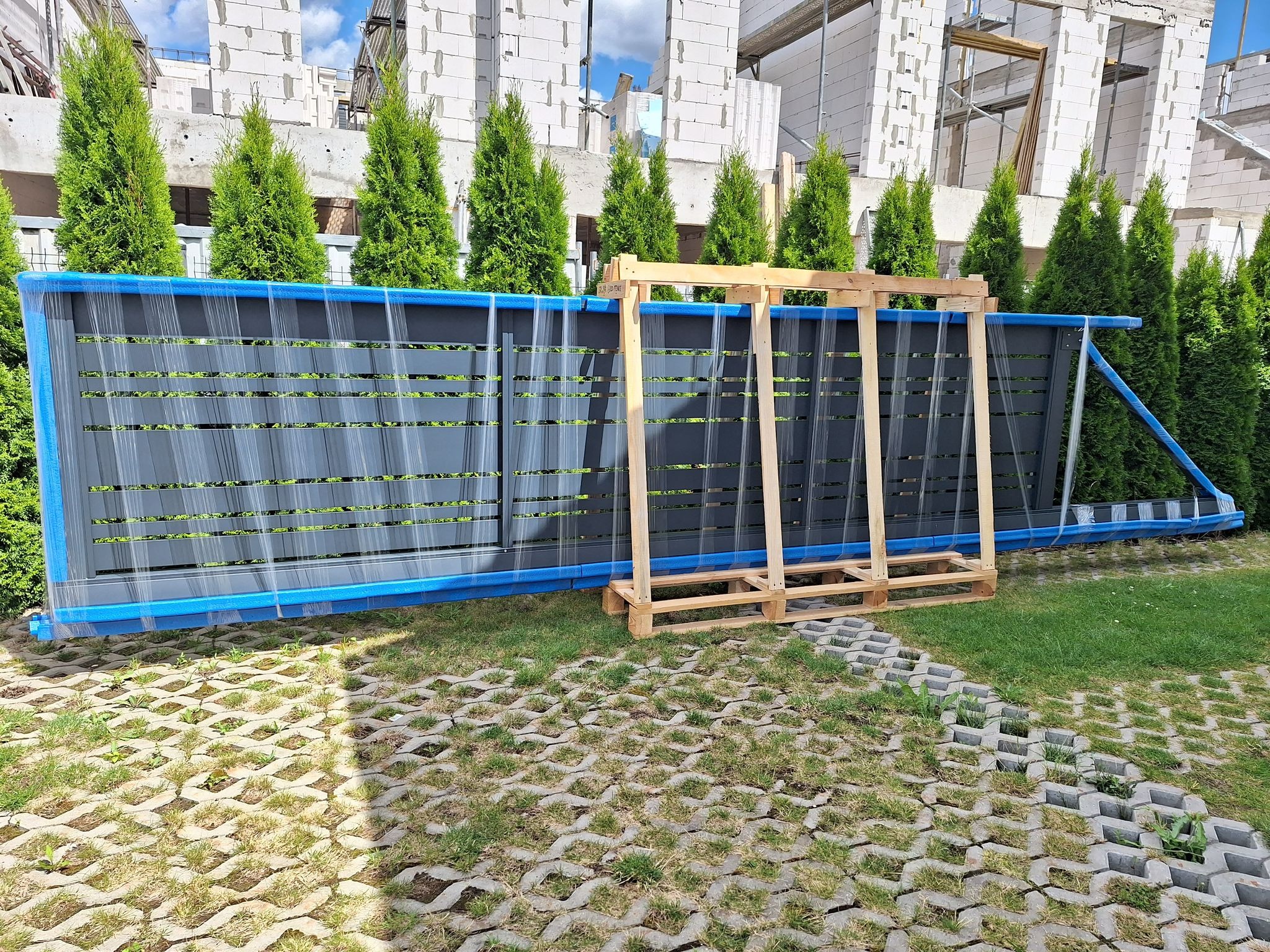 Szare panele ogrodzeniowe z niebieską ramą, zabezpieczone folią, oparte o drewnianą paletę na trawniku z ażurowych płyt, w tle rząd wysokich tuj i budynek w stanie surowym.