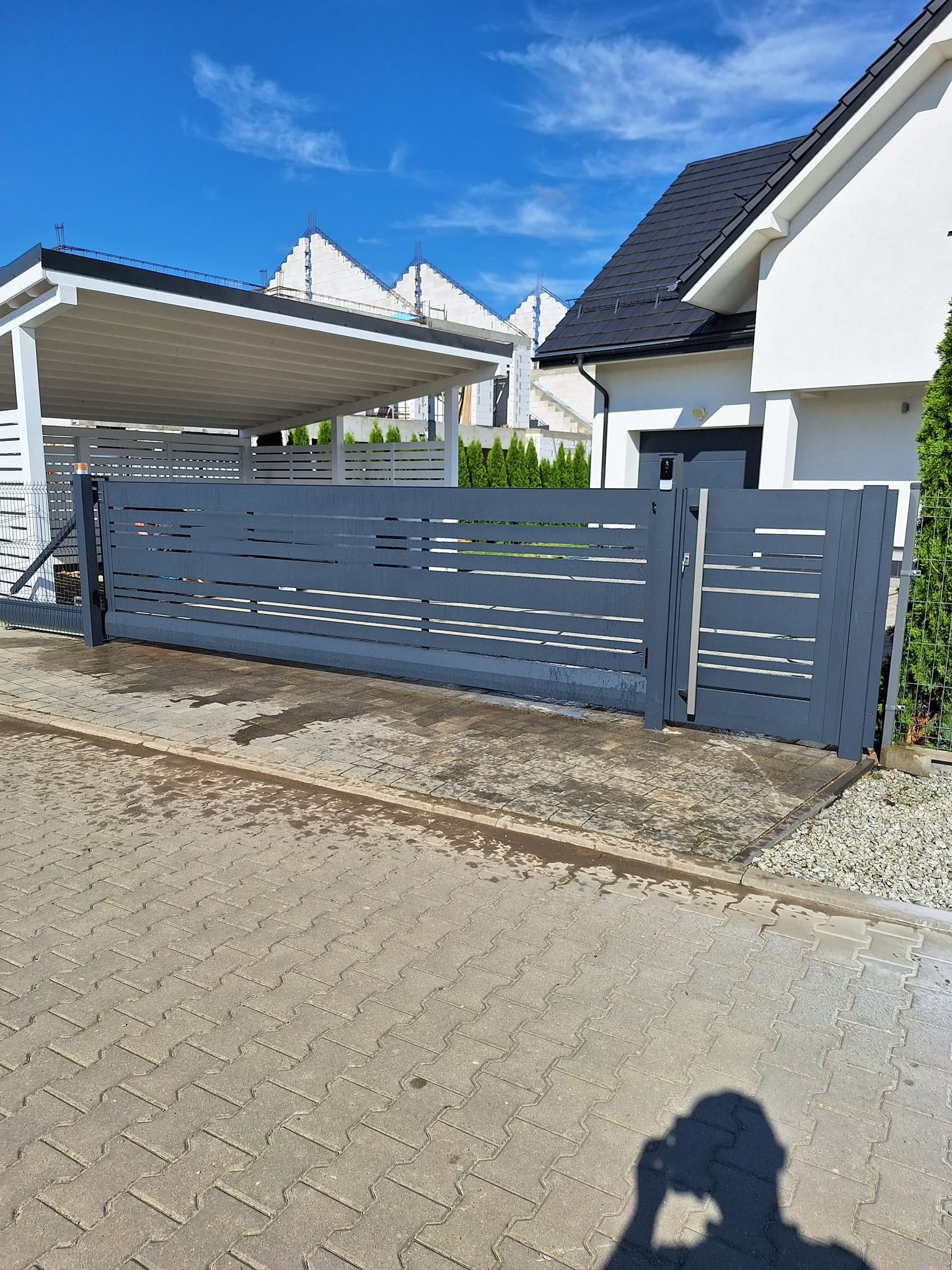 Przesuwna brama wjazdowa w kolorze grafitowym z poziomymi panelami, widoczna na tle nowoczesnego domu z podjazdem z kostki brukowej i wiatą garażową.