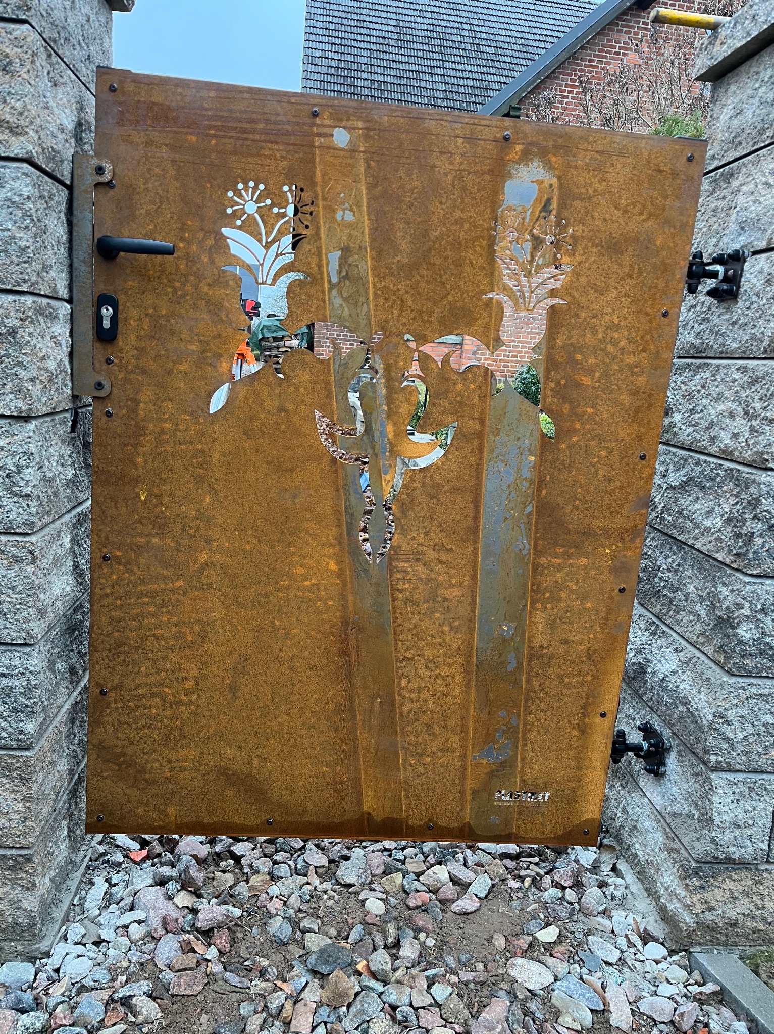 Rdzewiejąca metalowa brama z wyciętymi motywami roślinnymi, zamontowana między kamiennymi słupkami na tle domu z cegły i dachówki.