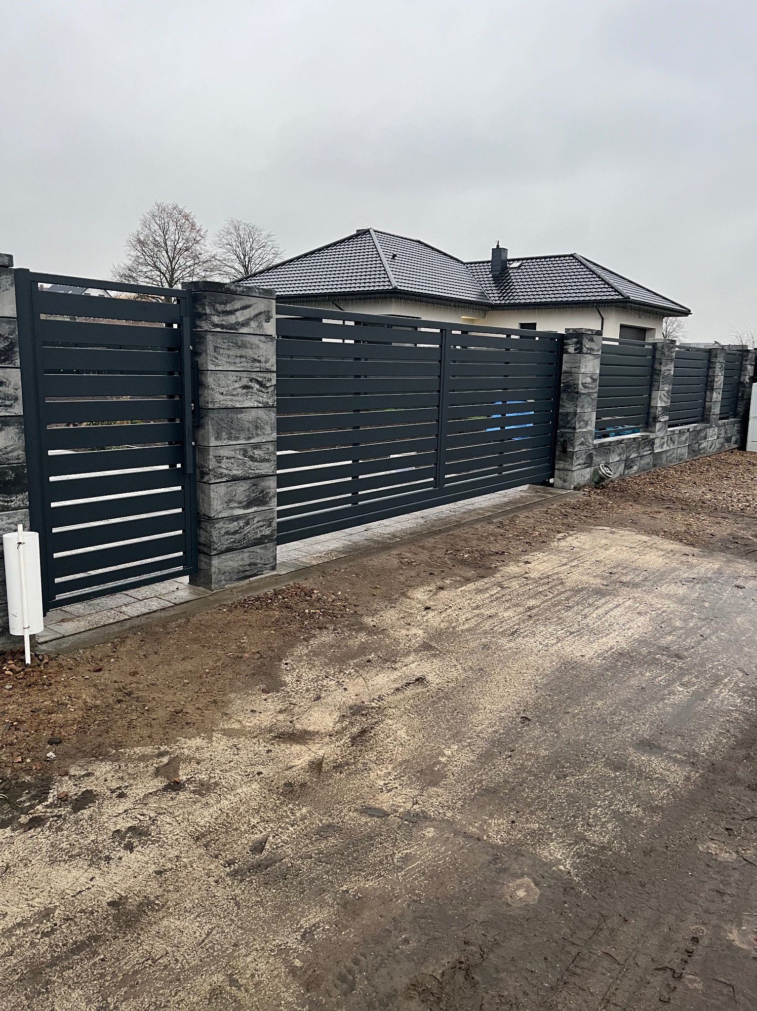 Nowoczesna brama przesuwna z poziomych, grafitowych paneli, osadzona w słupkach z szarego kamienia łupanego, na tle domu z ciemną dachówką i pochmurnego nieba.