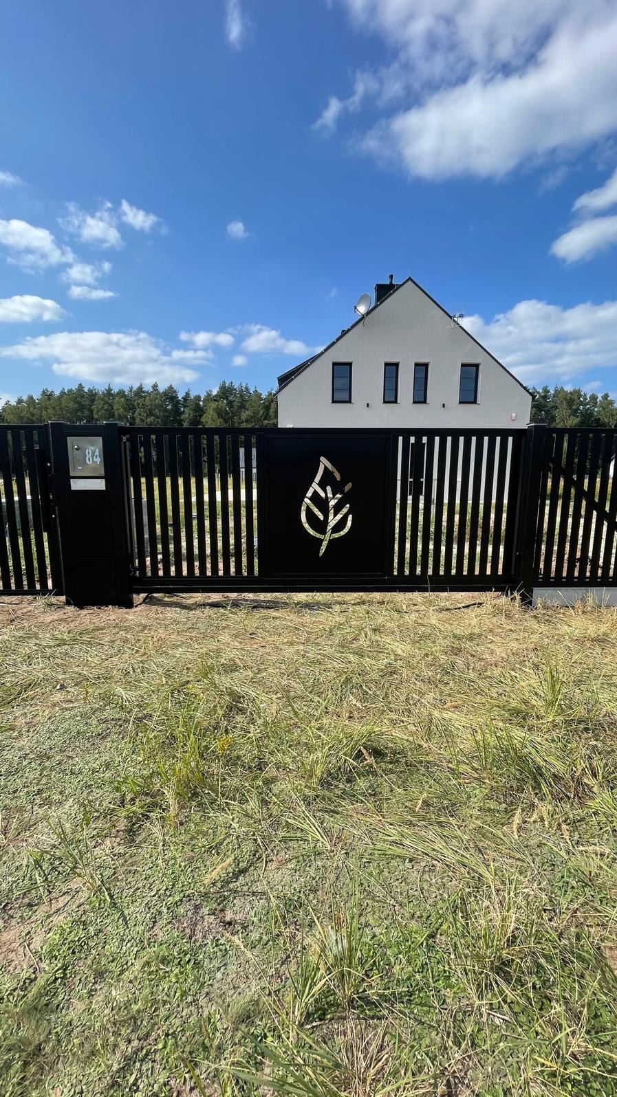 Czarna metalowa brama z motywem liścia, numer domu 84, nowoczesny dom w tle, trawa na pierwszym planie.