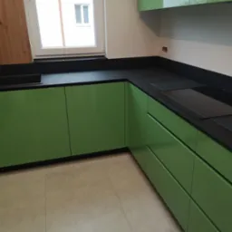 Nowoczesna kuchnia z zielonymi szafkami i czarnym blatem, widok na zlew i płytę grzewczą, jasna podłoga, okno z widokiem na zewnątrz.