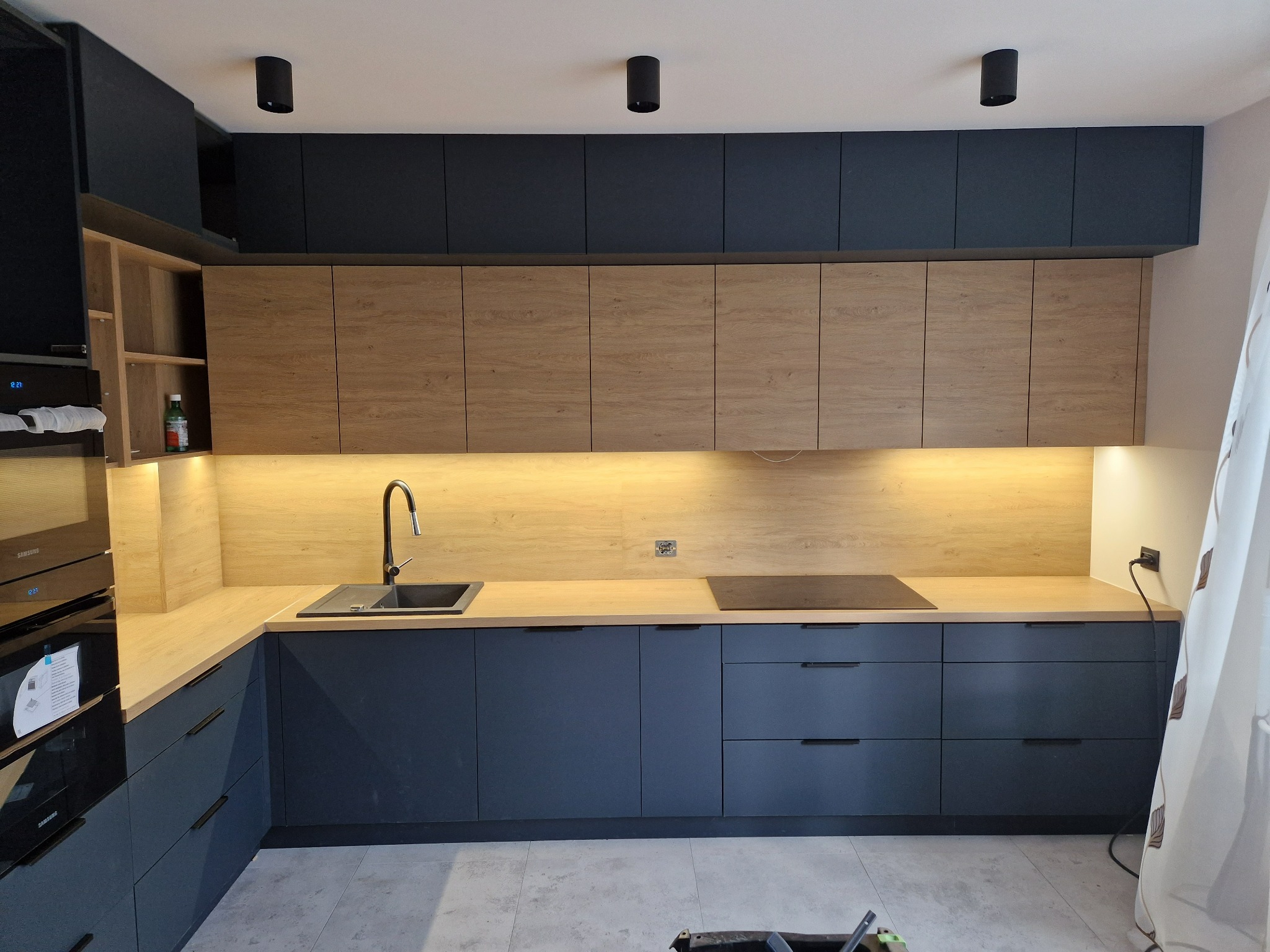 Nowoczesna kuchnia z drewnianymi i grafitowymi szafkami, podświetleniem LED pod górnymi szafkami i czarnymi punktowymi lampami sufitowymi.