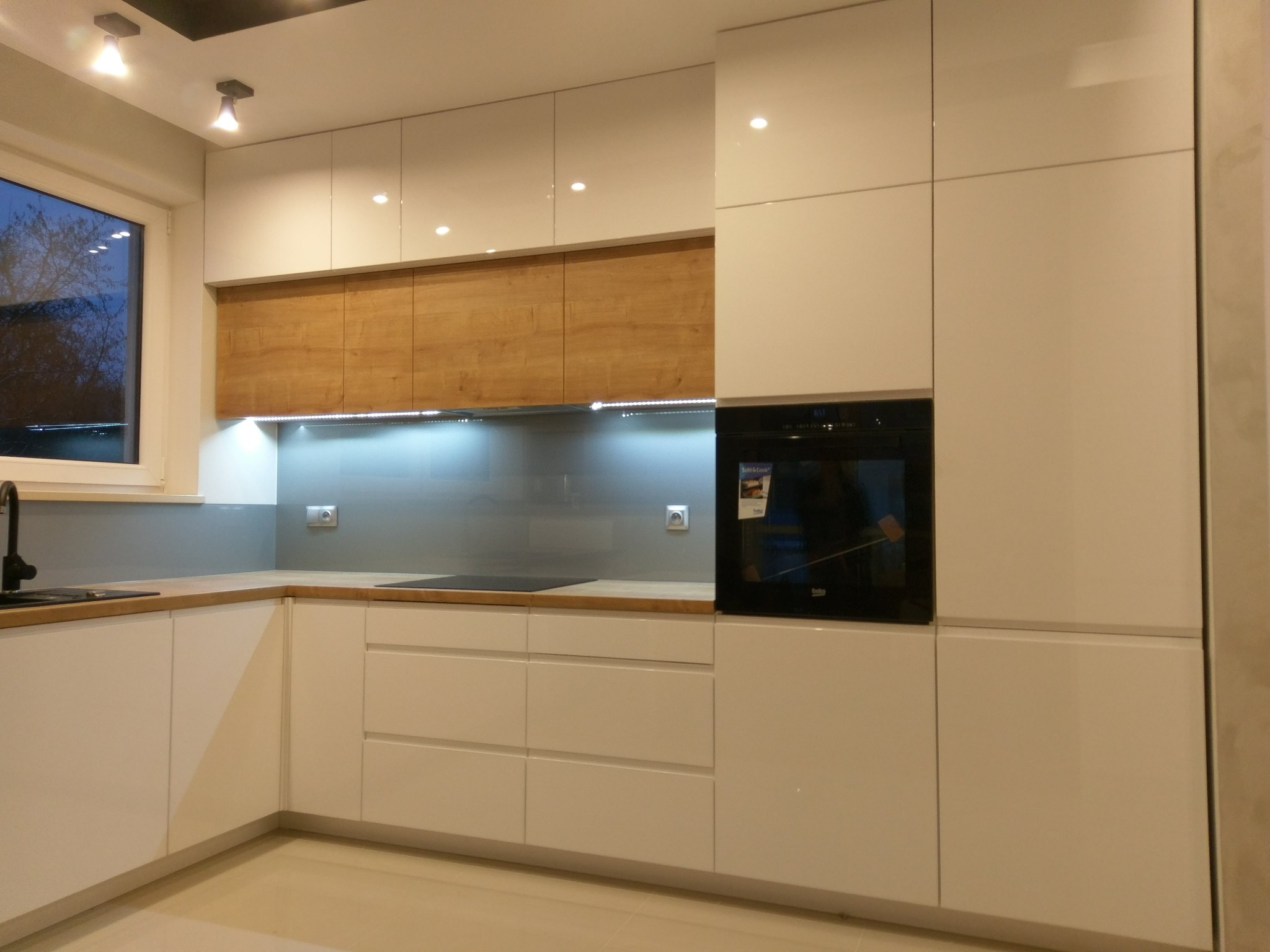 Nowoczesna kuchnia z białymi, lakierowanymi frontami szafek, drewnianym panelem nad blatem i wbudowanym piekarnikiem.