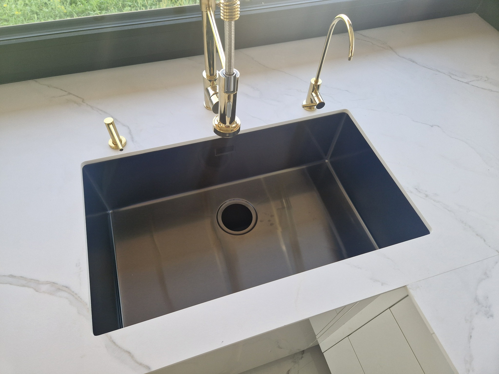 Czarny zlew wpuszczony w biały blat imitujący marmur, bateria i dozownik w kolorze złotym, widok z góry.