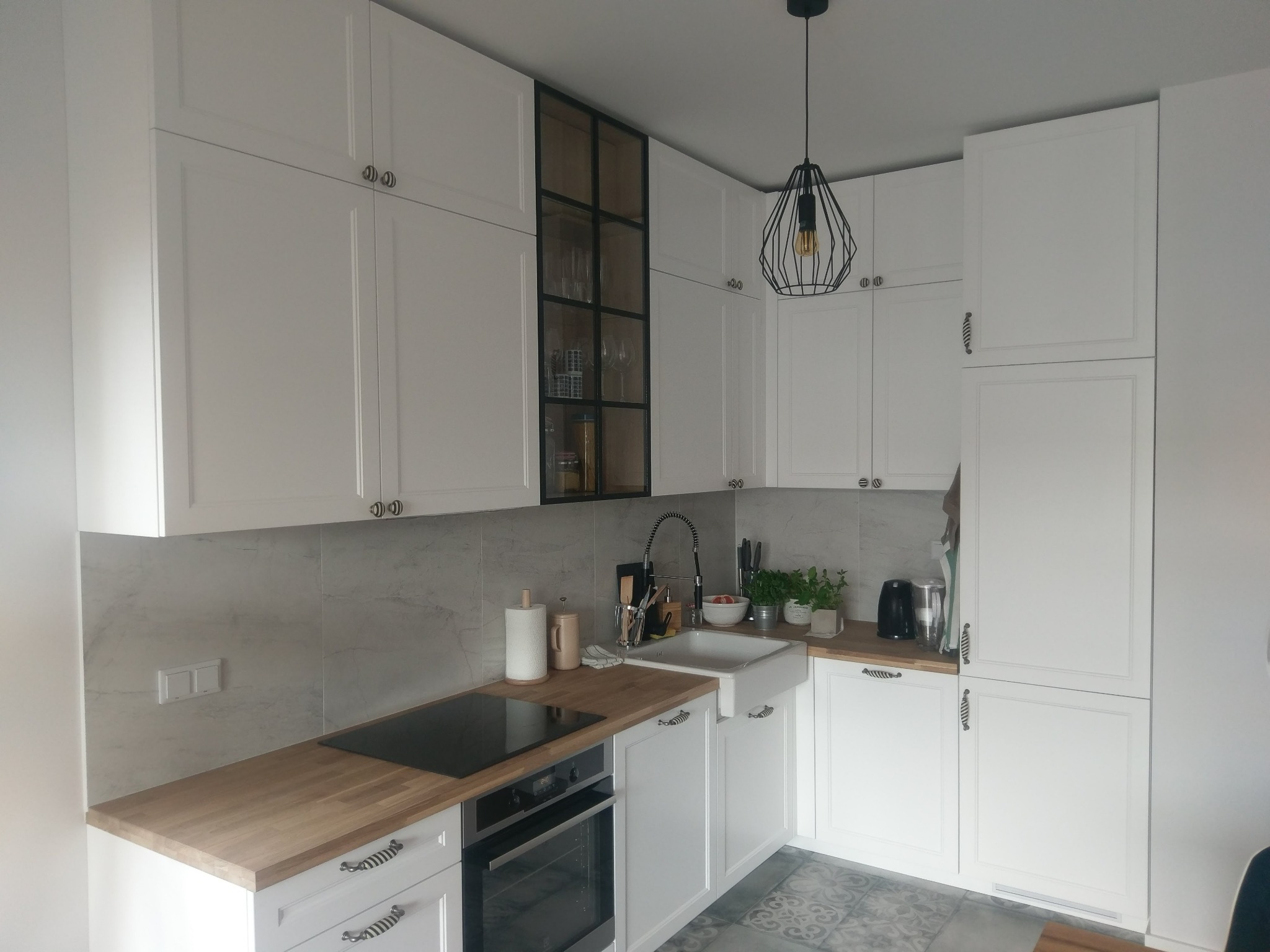 Jasna kuchnia z białymi szafkami z frezowanymi frontami, drewnianym blatem, ceramicznym zlewozmywakiem i czarną, geometryczną lampą wiszącą.