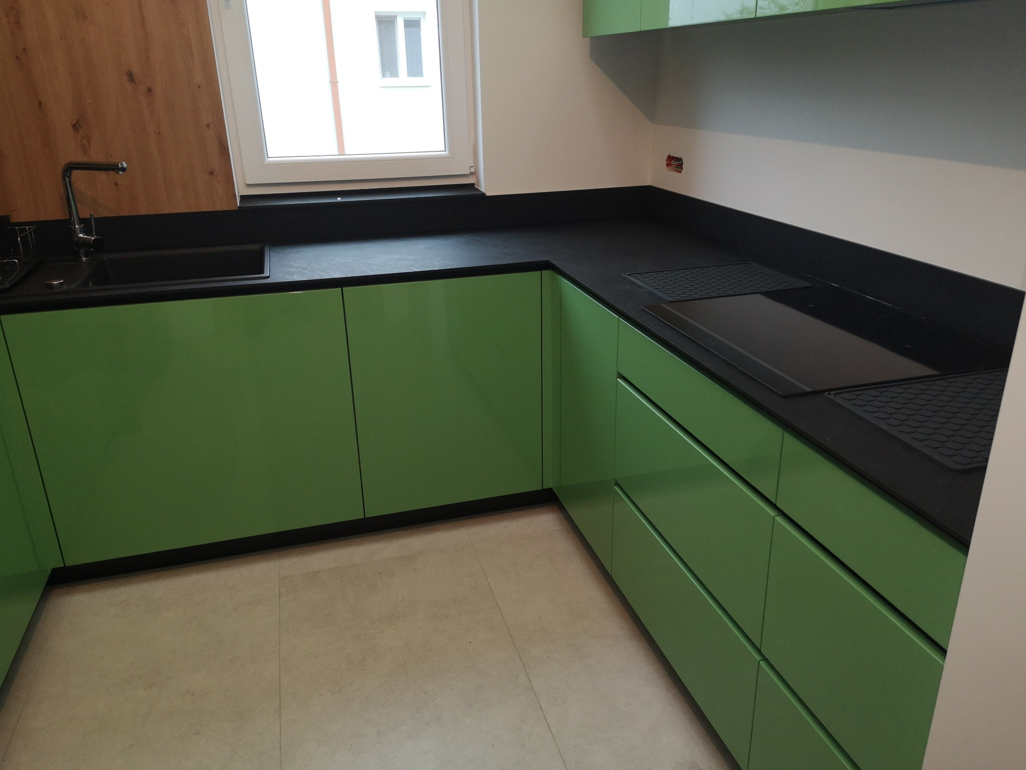 Nowoczesna kuchnia z zielonymi szafkami i czarnym blatem, widok na zlew i płytę grzewczą, jasna podłoga, okno z widokiem na zewnątrz.