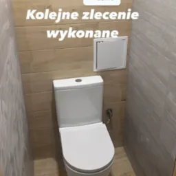 Biała toaleta kompaktowa w wykończonej łazience, widoczna klapka rewizyjna, ściany wyłożone płytkami imitującymi drewno i szary kamień.