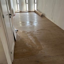 Horizon Piotr Kwiatkowski - Ułożona podłoga z płytek imitujących drewno w jasnym pomieszczeniu w trakcie prac wykończeniowych. Widoczne okna, drzwi i materiały budowlane.