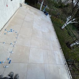 Horizon Piotr Kwiatkowski - Balkon z częściowo ułożonymi jasnymi, kwadratowymi płytkami, widoczne niebieskie krzyżyki dystansowe i stopy pracownika. W tle ogród z tulipanami.