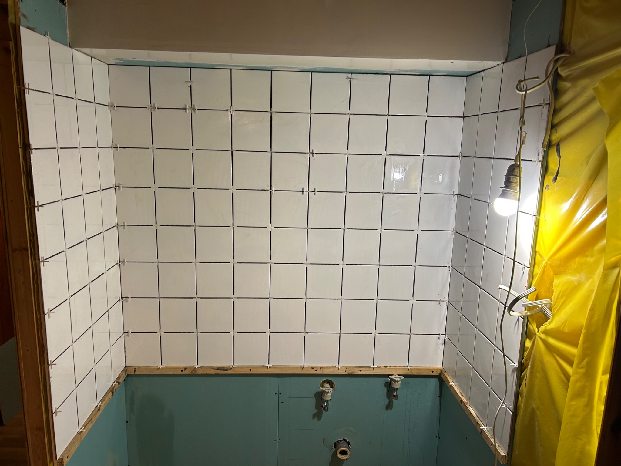 Ściany kabiny prysznicowej w trakcie układania białych, kwadratowych płytek z krzyżykami dystansowymi. Widoczne instalacje wodne i prowizoryczne oświetlenie.