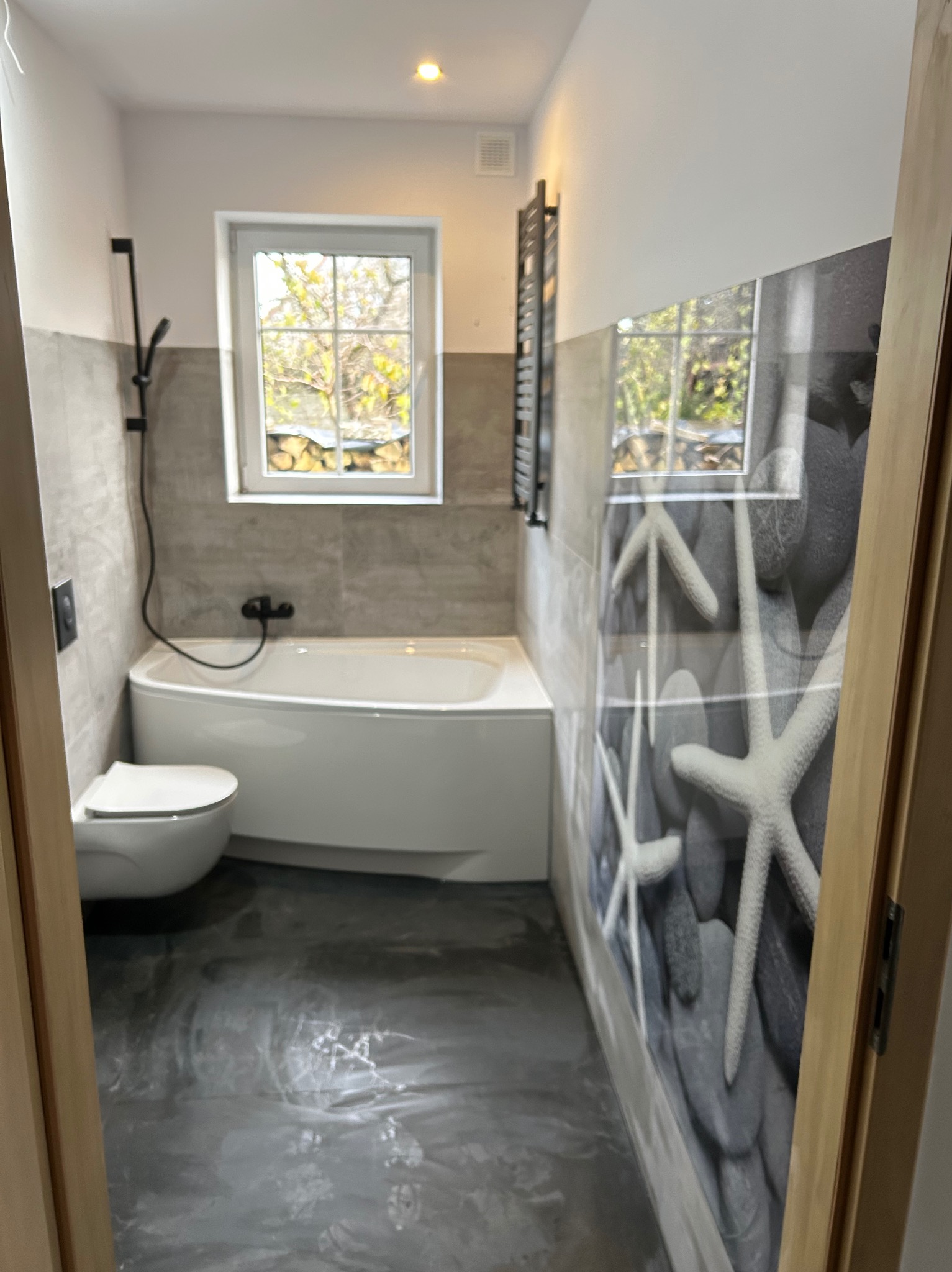 Wykończona łazienka z wanną, toaletą i grzejnikiem drabinkowym. Na ścianie dekoracja z motywem kamieni i rozgwiazd. Podłoga w odcieniach szarości.