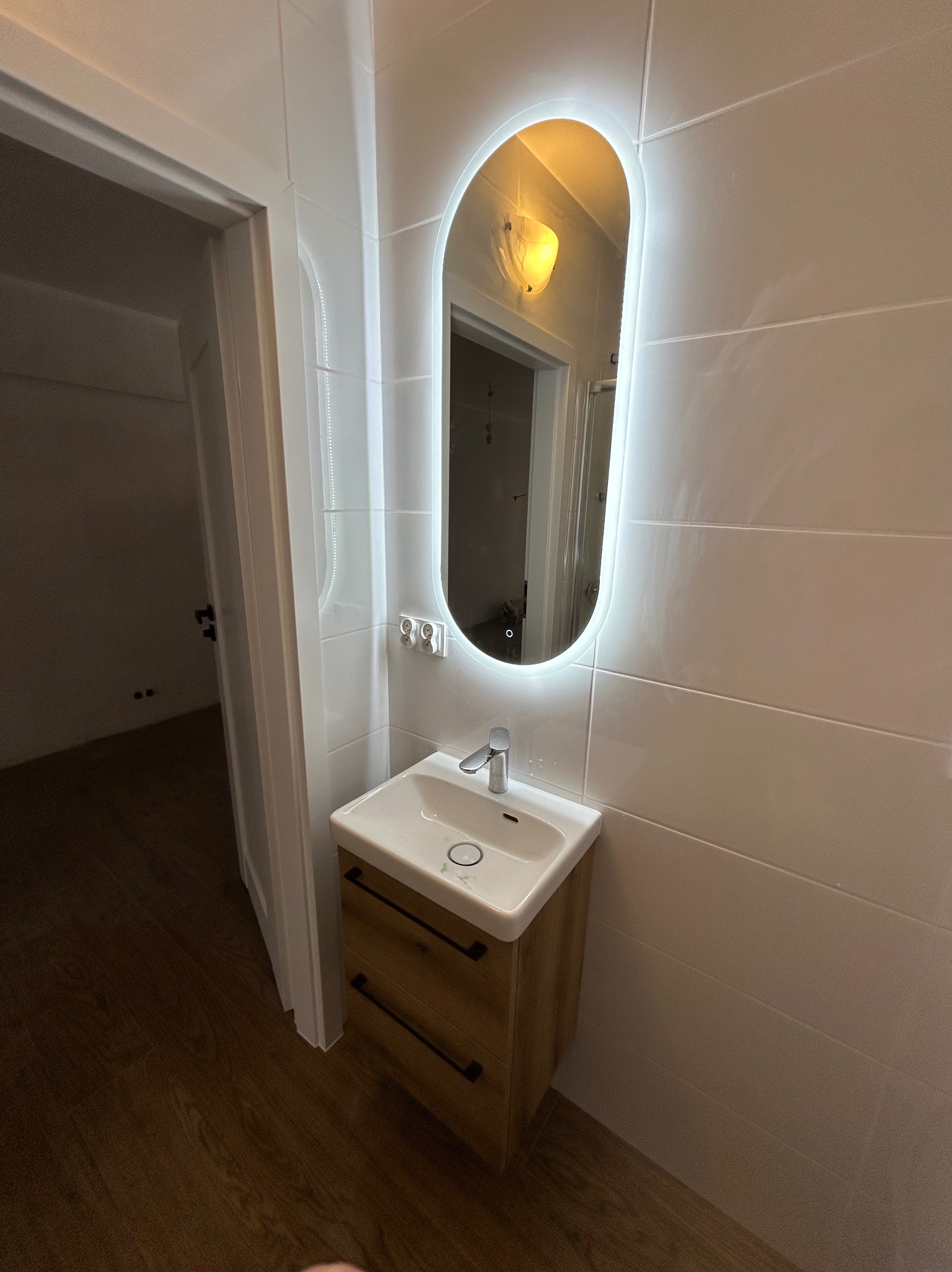 Biała łazienka z owalnym lustrem LED, drewnianą szafką pod umywalką i białymi płytkami. Widoczny fragment podłogi z paneli i otwarte drzwi do innego pomieszczenia.