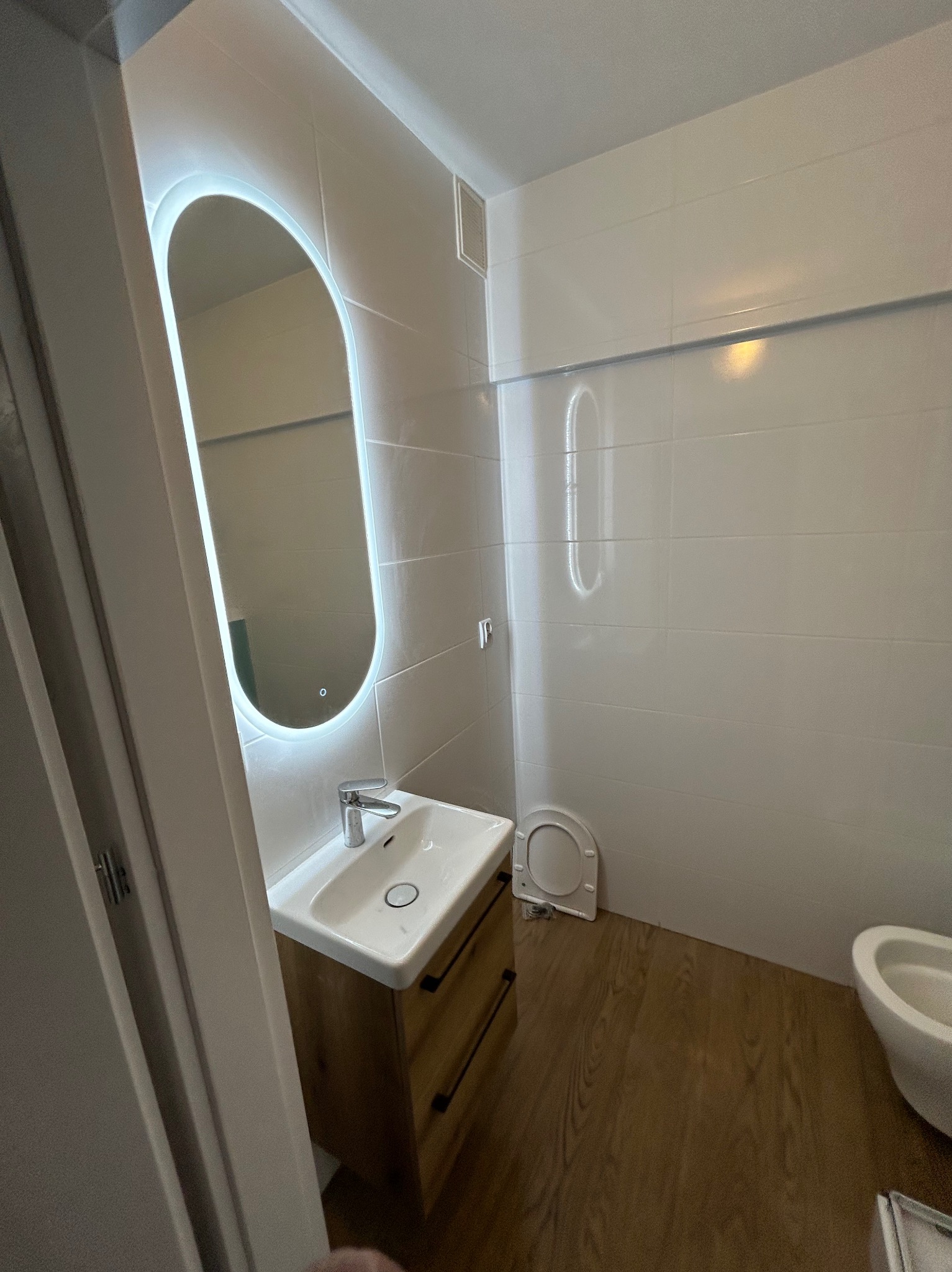 Nowoczesna łazienka z podświetlanym lustrem o zaokrąglonym kształcie, drewnianą szafką pod umywalką i białymi płytkami na ścianach. Widoczny fragment toalety i drewniana podłoga.