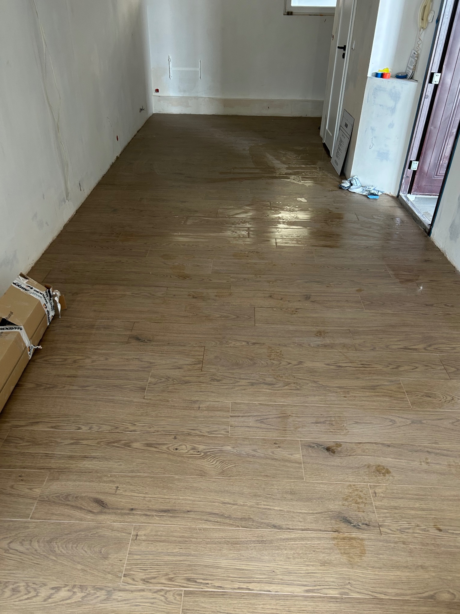 Ułożona podłoga z płytek imitujących drewno w jasnym odcieniu, widoczne kartony z płytkami przy ścianie, surowe ściany przygotowane do dalszych prac wykończeniowych.